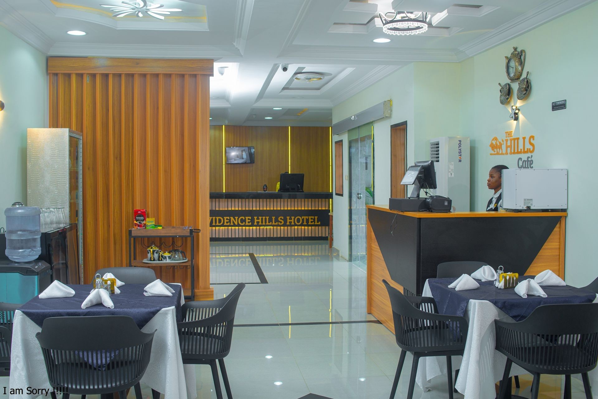 Hotel reception area with a desk, a staff member, water dispenser, and dining table setup.