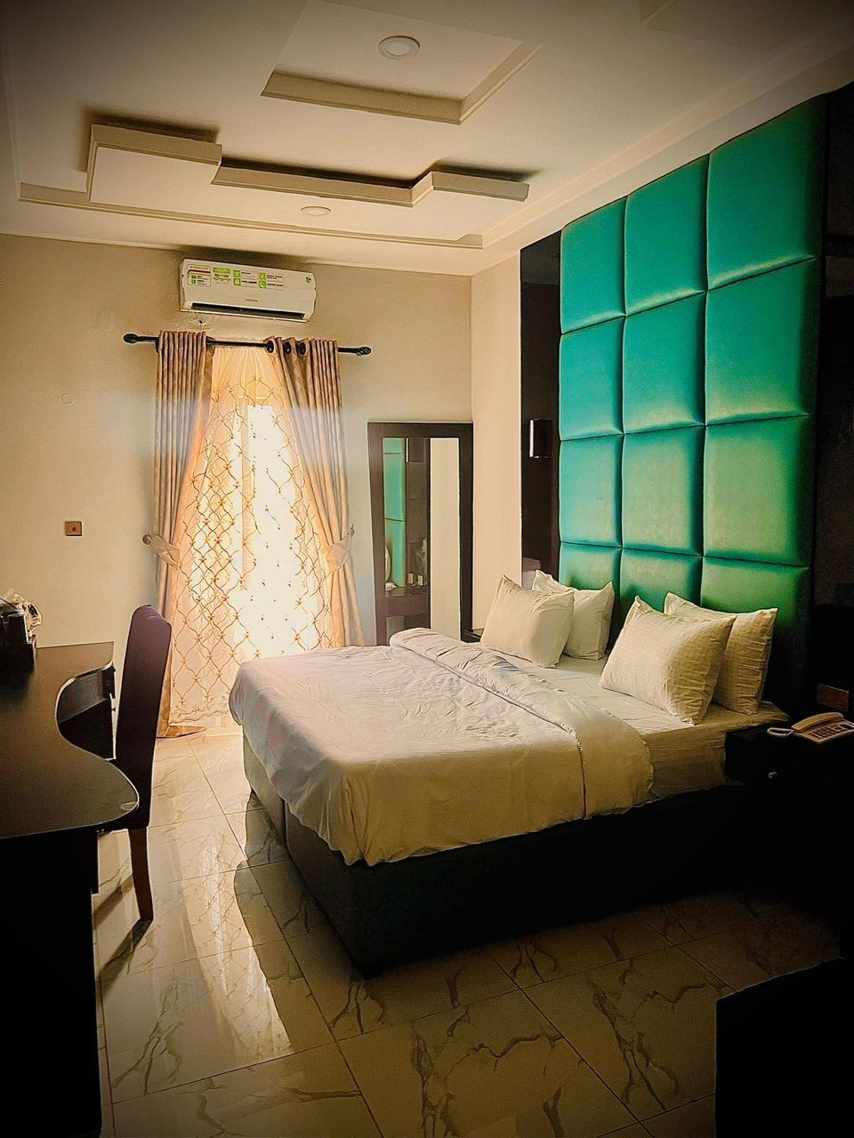 Modern hotel room with a large bed, teal padded headboard, and sunlight streaming through curtains.