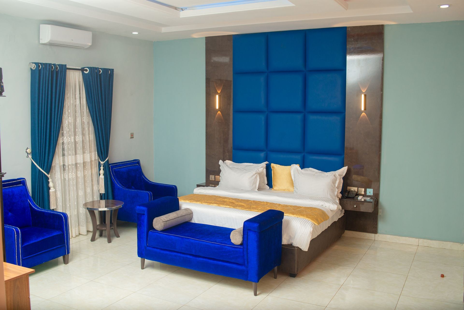 Modern bedroom with a large blue padded headboard, matching blue furniture, and warm lighting.